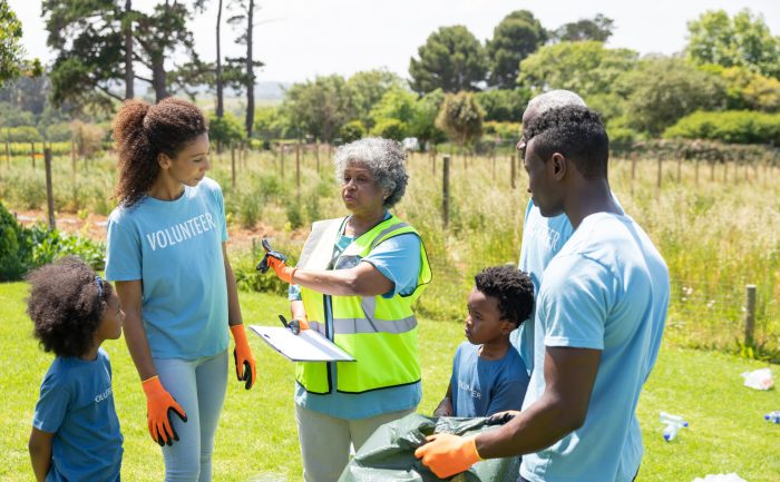 Front view of a senior African American woman standing in a field wearing a hi-vis vest and gloves, holding a clipboard and instructing a diverse group of African American volunteers before collecting rubbish and recycling